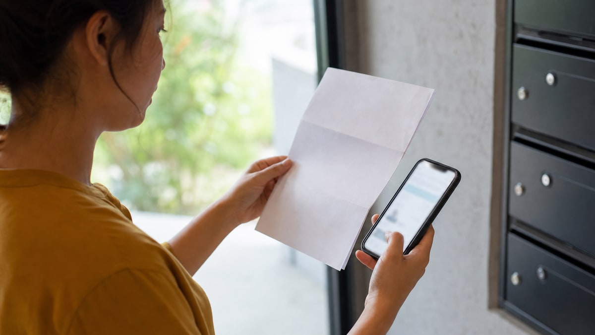 A person checking a phone while holding a credit account notice near apartment mailboxes.